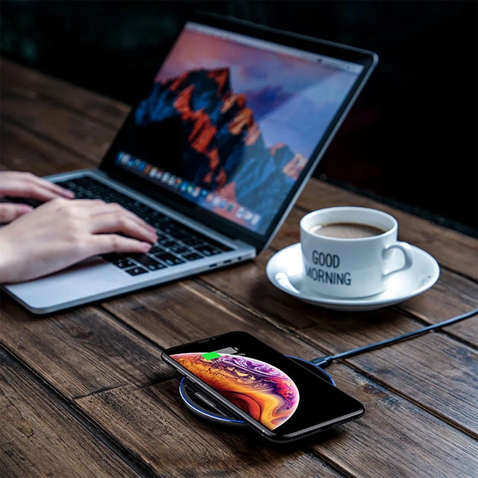 Person using a laptop with a smartphone on a wireless charger, and a cup of coffee on a wooden table.